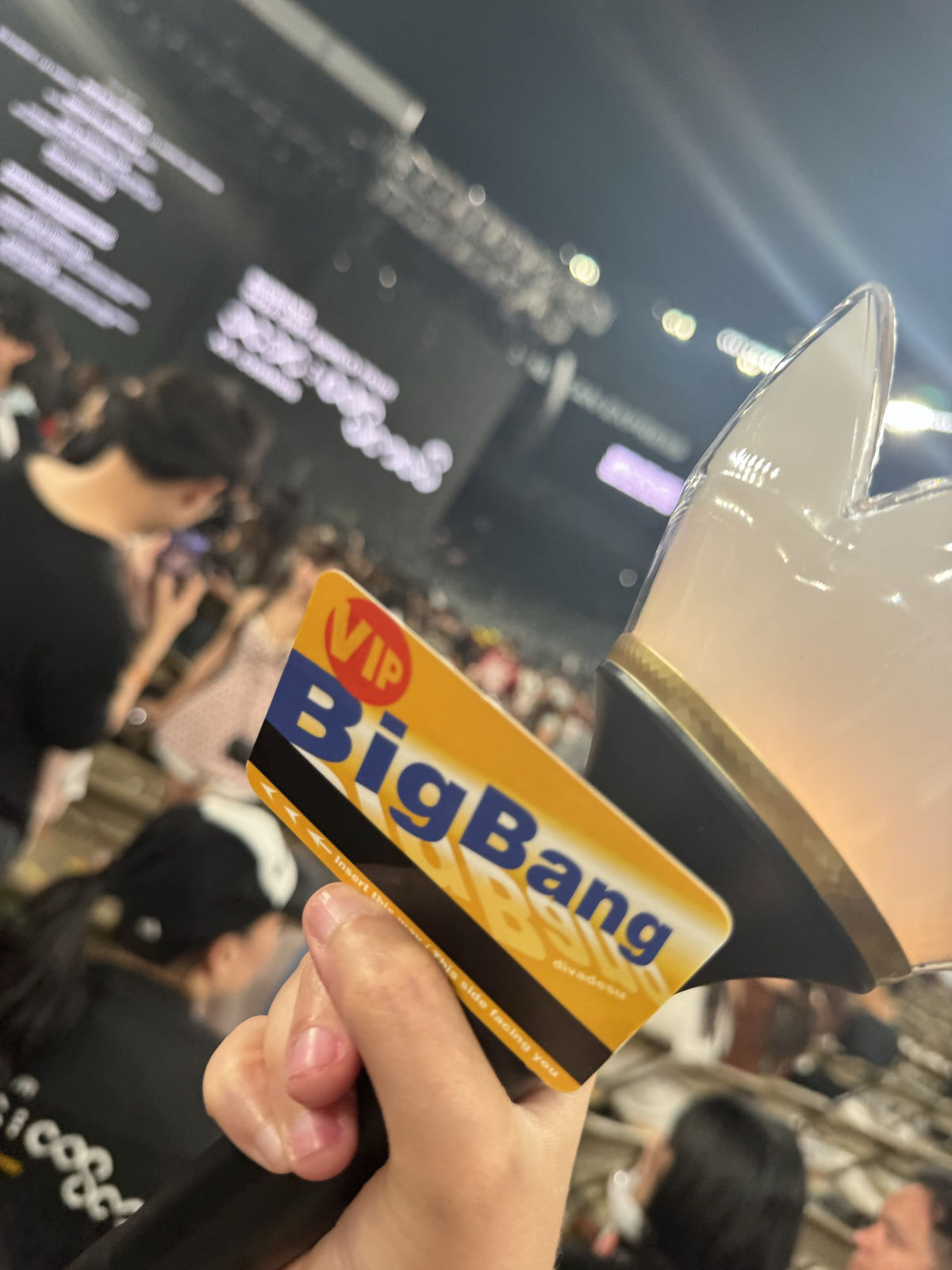 A hand holds a Big Bang VIP light stick and fanmade card in a crowded indoor event space. The card features a yellow and blue 'VIP Big Bang' label