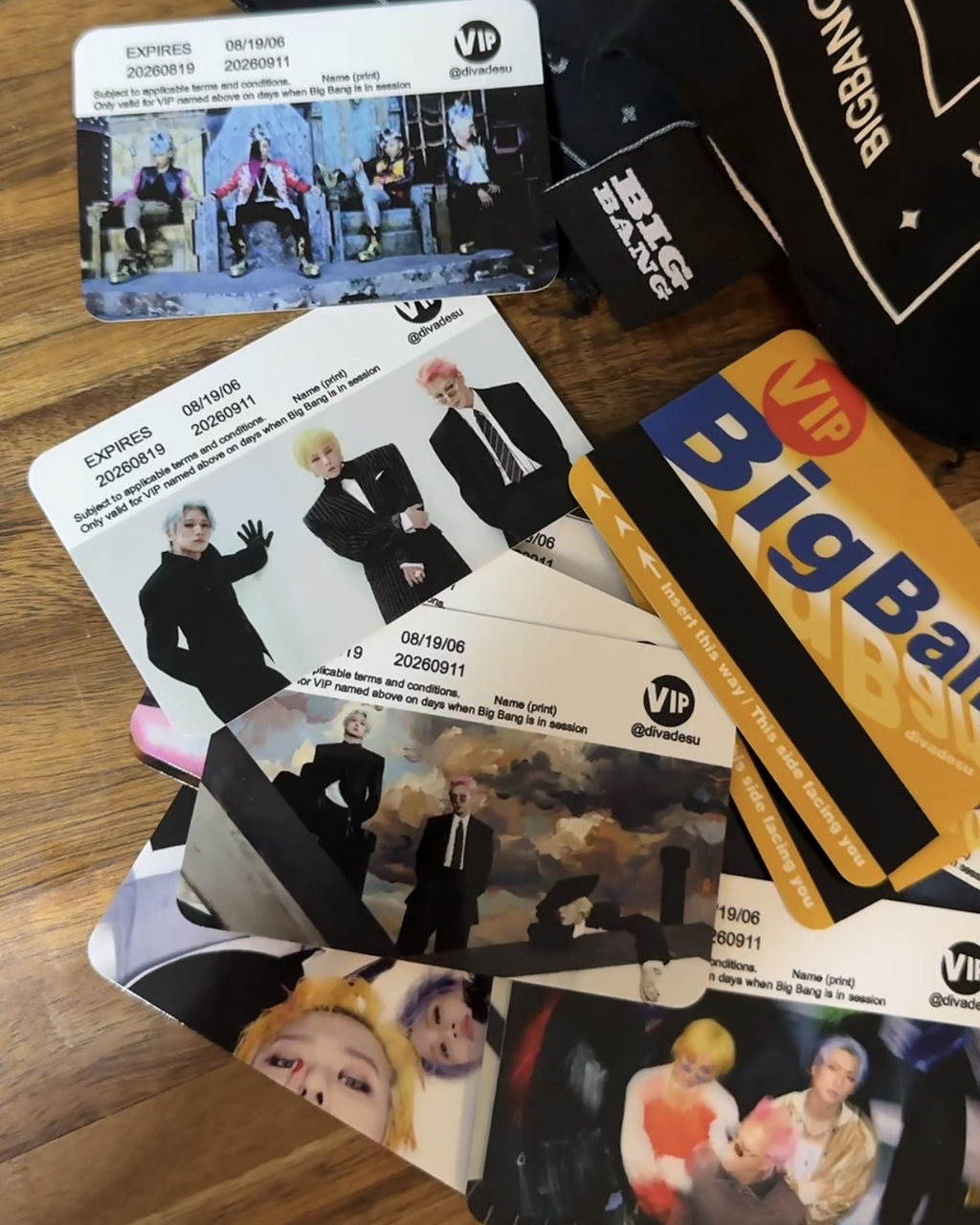 A pile of printed fanmade VIP card-style passes and a black cap with white text laid on a wooden surface. The cards feature photos of people in suits and the word 'BigBang' with a circle VIP logo.