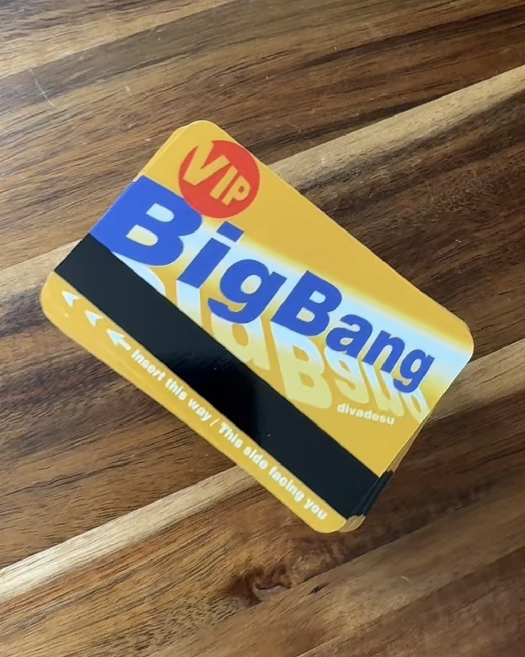 A yellow and blue fanmade Big Bang VIP transit card with a black magnetic stripe, resting on a wooden surface.
